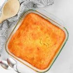 A full glass baking dish of baked Jiffy corn pudding with spoons and ladle on the side.