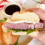 A close-up of a hand holding a prime rib sandwich with a toasted bun, filled with creamy sauce.