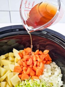 A measuring cup pouring broth over diced vegetables in a slow cooker.