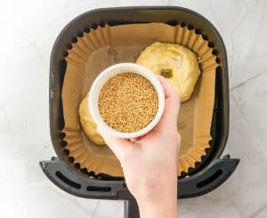 Hand holding sesame seeds over bagels in the air fryer basket.