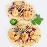 Two shrimp tacos topped with red cabbage, onions, and sauce on tortillas, garnished with cilantro and jalape&ntilde;o.