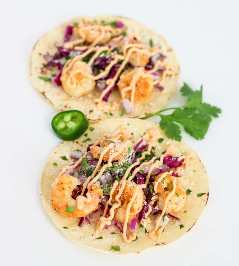 Two shrimp tacos topped with red cabbage, onions, and sauce on tortillas, garnished with cilantro and jalapeño.