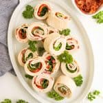 Overhead view of turkey clubhouse pinwheels arranged on a platter with fresh parsley and a small bowl of crumbled bacon.