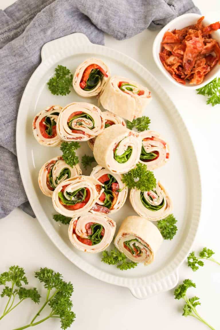Overhead view of turkey clubhouse pinwheels arranged on a platter with fresh parsley and a small bowl of crumbled bacon.