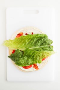 Romaine lettuce leaves are added over the tortilla with cream cheese mixture, crushed bacons, provolone cheese, deli turkey, and tomatoes.