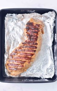 Grilled pork tenderloin transferred on foil in a baking pan.