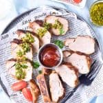 Overhead shot of a full serving tray with grilled pork slices, chimichurri, and tomatoes, alongside a small bowl of whole cherry tomatoes.