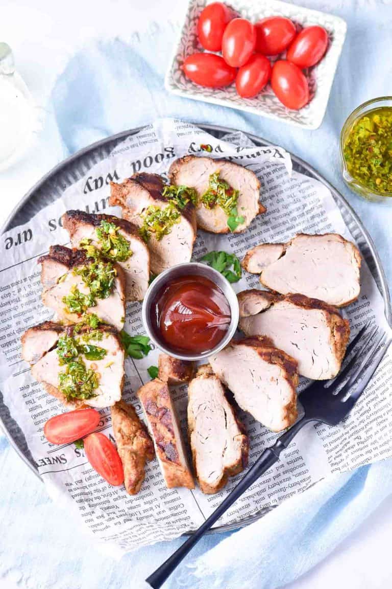 Overhead shot of a full serving tray with grilled pork slices, chimichurri, and tomatoes, alongside a small bowl of whole cherry tomatoes.