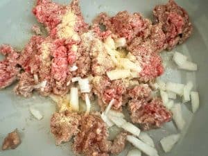 Raw ground beef with chopped onion and seasoning in a pan.
