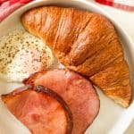 Overhead view of plate with croissant, fried egg, ham slices, and coffee.