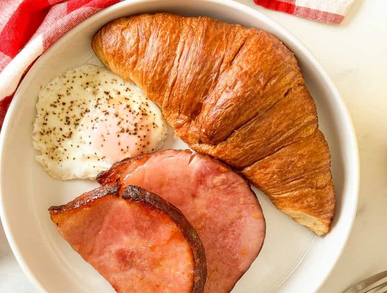 Overhead view of plate with croissant, fried egg, ham slices, and coffee.
