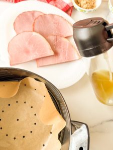Sliced ham steaks on a white plate next to an air fryer basket lined with parchment paper.