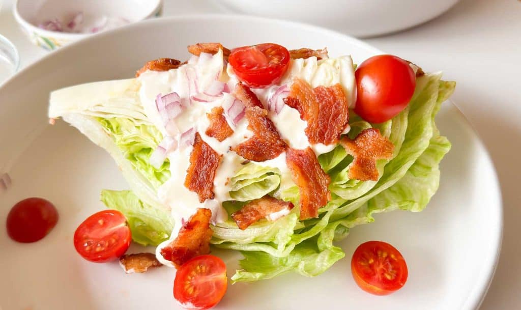 Lettuce wedge topped with dressing, red onion, cherry tomatoes, and crispy bacon pieces on a white plate.