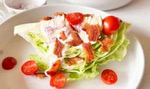 Lettuce wedge topped with dressing, red onion, cherry tomatoes, and crispy bacon pieces on a white plate.