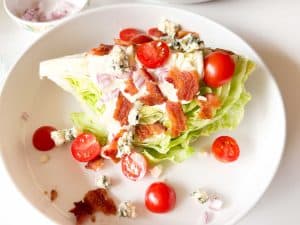 Overhead view of a wedge salad with dressing, bacon, tomatoes, red onion, and blue cheese on a white plate.