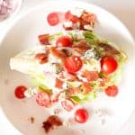 Wedge salad on a plate with small bowls of dressing, bacon, blue cheese, and chopped onion nearby.