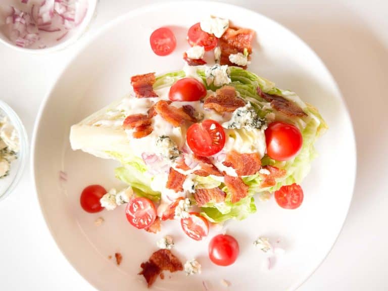 Wedge salad on a plate with small bowls of dressing, bacon, blue cheese, and chopped onion nearby.