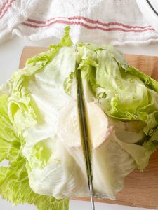 Head of iceberg lettuce cut in half on a cutting board with a knife down the center, kitchen towel in the background.