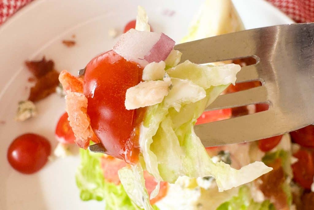 Close-up of a fork holding a bite of lettuce, tomato, bacon, and blue cheese from a wedge salad.