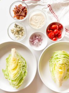 Ingredients arranged for wedge salad: lettuce wedges, cherry tomatoes, chopped onion, crumbled blue cheese, bacon pieces, and dressing in small bowls.