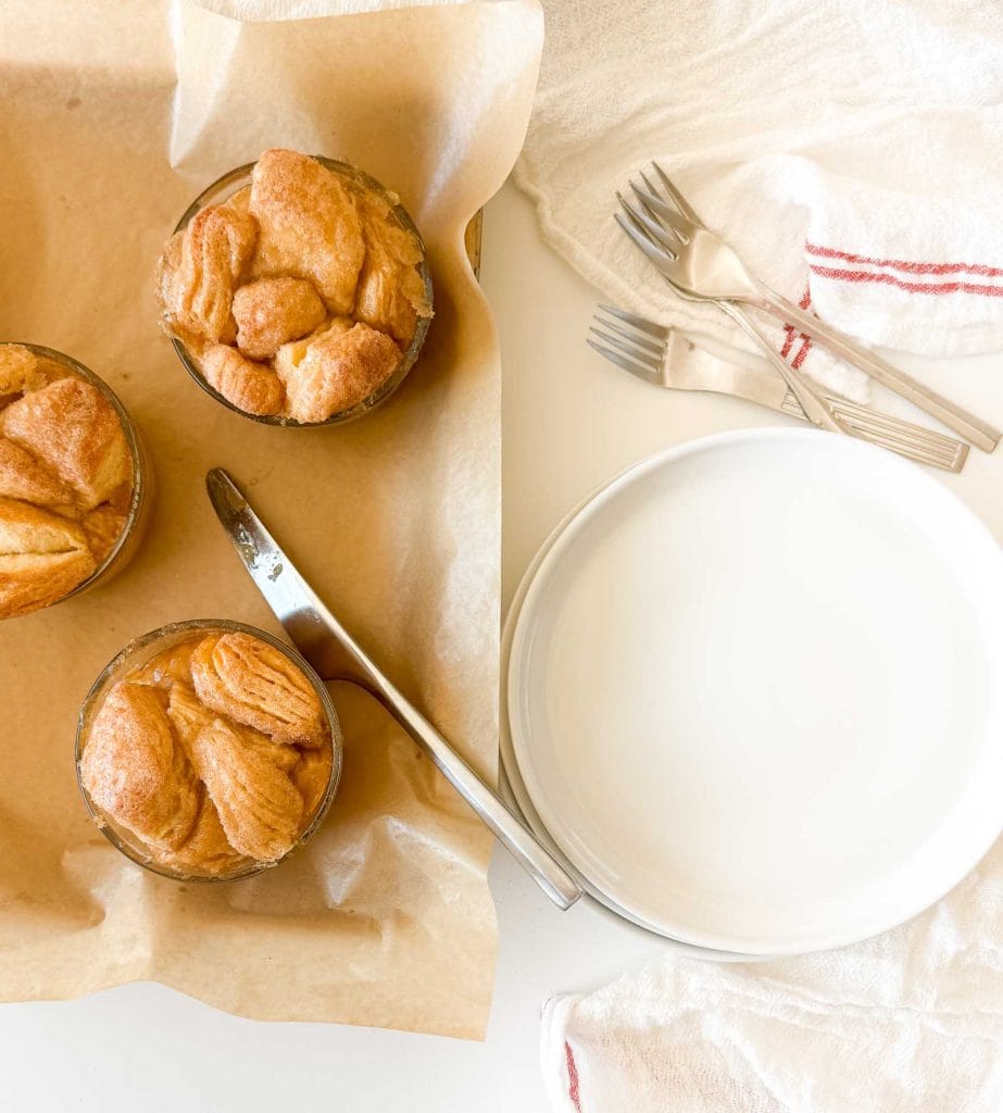Three small monkey bread portions in glass dishes on parchment paper beside a stack of white plates and forks.