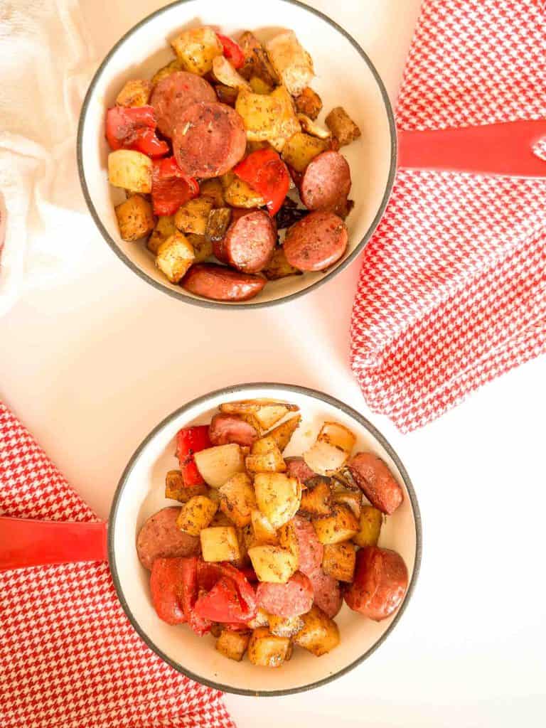 Two bowls filled with roasted sausage, potatoes, onions, and red bell peppers on a light surface.