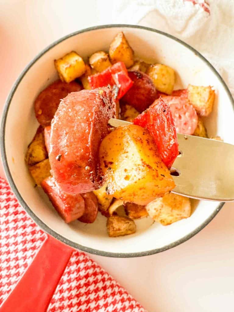 Close-up of a fork holding a piece of sausage, roasted potato, and red pepper over a bowl.
