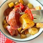 Close-up of fork lifting sausage, potato, and pepper from a bowl of roasted vegetables.