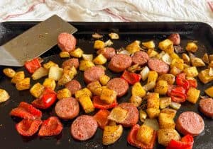 Roasted sausage, potatoes, and peppers on a sheet pan with a spatula nearby.