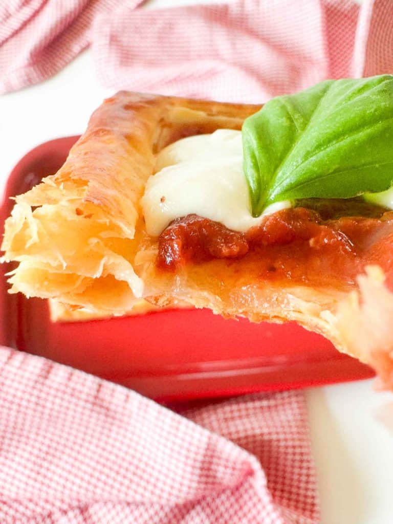 Bite taken from a puff pastry pizza showing flaky layers, pizza sauce, melted mozzarella, and basil.