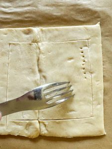 Fork poking holes in the center of puff pastry rectangles with scored edges on parchment paper.