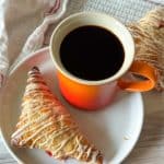 Strawberry turnover on a plate next to an orange mug of coffee, with another pastry on a cooling rack behind.
