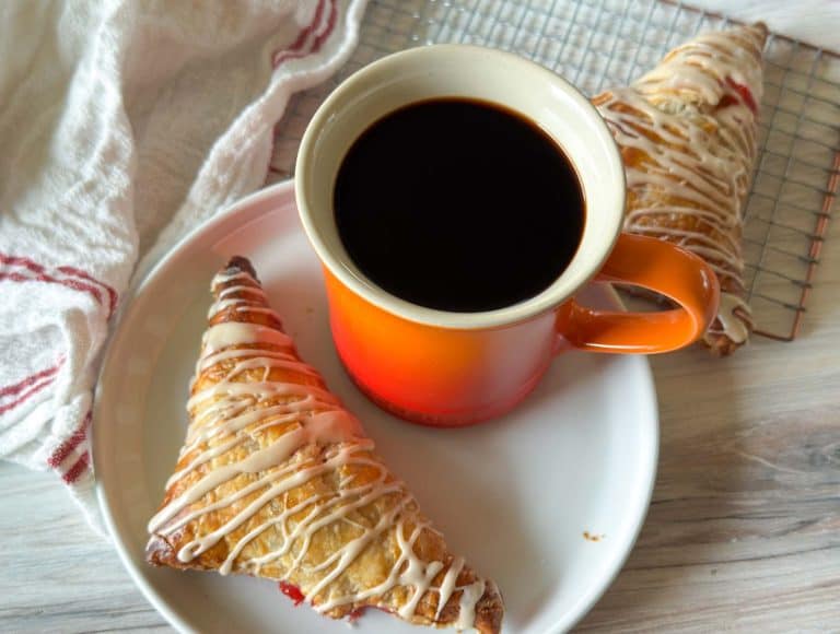 Strawberry turnover on a plate next to an orange mug of coffee, with another pastry on a cooling rack behind.